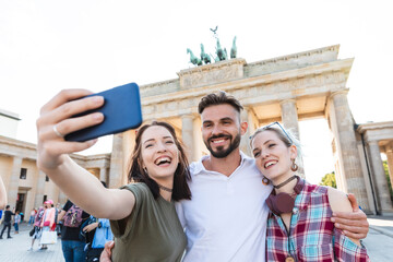 Portrait of three happy friends taking selfie with cell phone in front of Brandenburger Tor,...