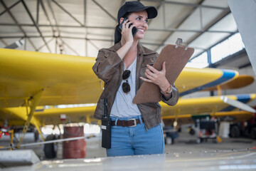 Female pilot talking on the phone in hangar - obrazy, fototapety, plakaty