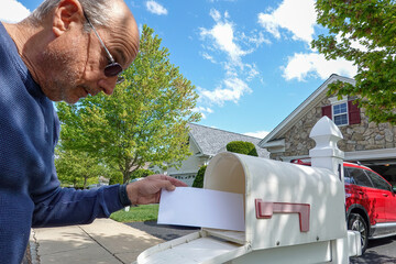 Older bald senior citizen man looks at a white envelope as he takes it out of letterbox