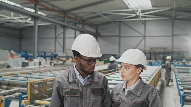 Two mixed-raced engineers making decision or discussing breakdown of production machines at industrial factory. People considering constructions and communicating in plant. Servicing technology.