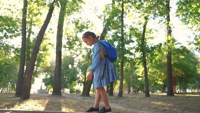 little girl walking along the curb. concept of a happy childhood and loving family. a schoolgirl in a dress and with a blue backpack walks along the lifestyle curb, keeps her balance