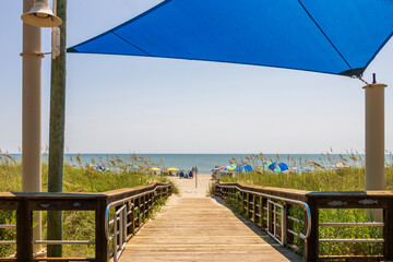 A beautiful summer landscape at the beach with a wooden footpath, people relaxing on the beach with colorful umbrellas, blue ocean water and blue sky in Carolina Beach North Carolina USA