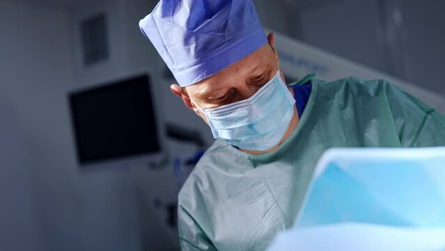 Mid-aged male surgeon working in modern surgery room. Male anesthesiologist walks at backdrop.