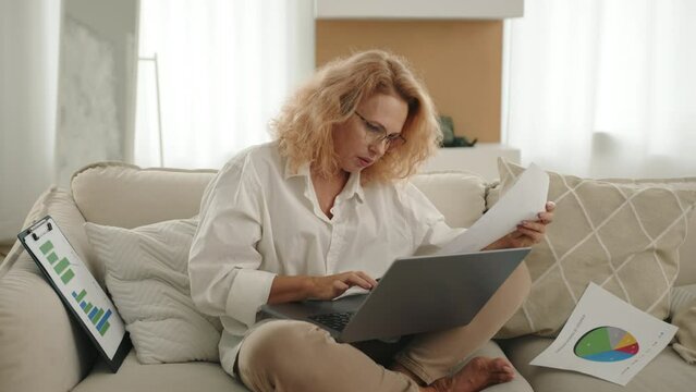 Pretty businesswoman in glasses working from home, sitting in a cozy room in yoga pose inputting data into laptop, holding shit of paper, checking document. High quality 4k footage
