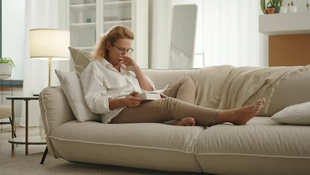 Nice mature blonde in glasses yawning, putting book aside on sofa, turning to her side, closing eyes and peacefuly falling asleep at daytime in modern flat. High quality 4k footage
