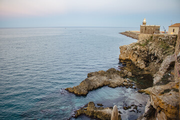 Vista de un acantilado rocoso junto al mar, con un faro y edificaciones en la costa durante el atardecer. Ideal para capturar la majestuosidad y serenidad de un entorno costero.