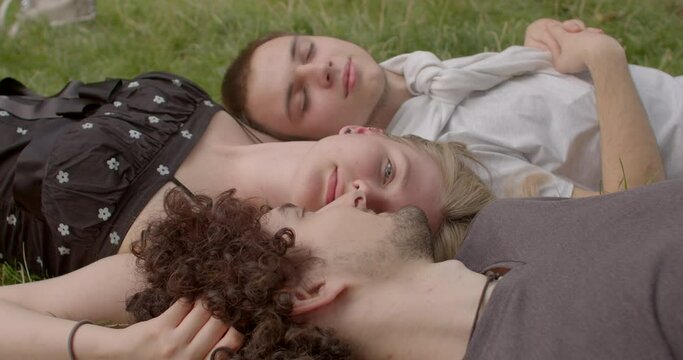 Girl positioned between two guys, tenderly kissing one of them. Love triangles as intricacies of human emotions. Lifestyle portraits of students after a lecture, young people lying on the grass. 