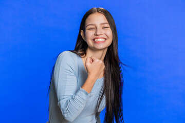 Fototapeta premium Happy joyful woman celebrating success victory, winning birthday, lottery jackpot goal achievement play game good positive news, triumph. Young smiling brunette girl on blue studio wall background