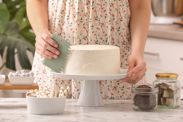 Woman using scraper to decorate cake at white marble table in kitchen, closeup