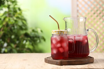 Refreshing hibiscus tea with ice cubes on wooden table against blurred green background. Space for text