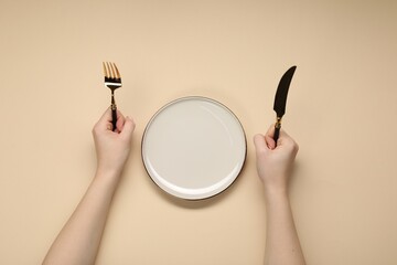 Woman holding fork and knife near empty plate at beige table, top view