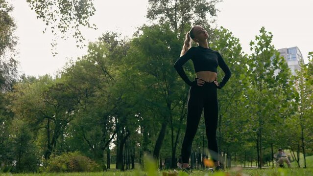 A sporty girl stretches her shoulders and neck during morning exercises in park while listening to music on headphones