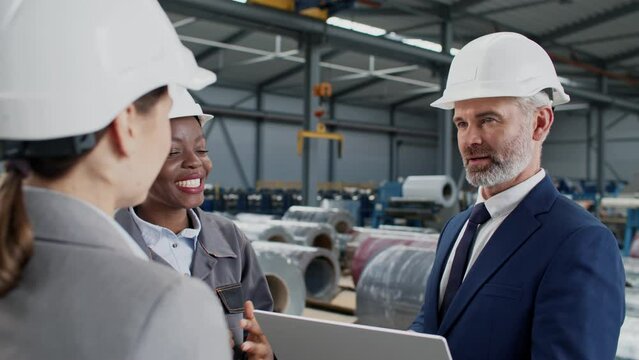 Mixed-raced people with helmets having meeting in storage at large factory. Charming forewoman telling about results of manufacturing in facility. Manager actively listening while working on laptop.