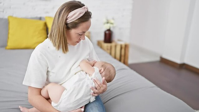 Devoted mother sitting on a relaxed bedroom bed indoors, showing utmost concentration while lovingly breastfeeding her infant. cherishing the athletic home-nurturing lifestyle with seriousness.