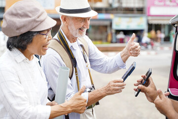 Obraz premium Closeup American senior tourist man poses happy thumbs up and paying the fare to the Tuktuk Thailand taxi driver by smart phone on blurred of city background. Senior tourist concept