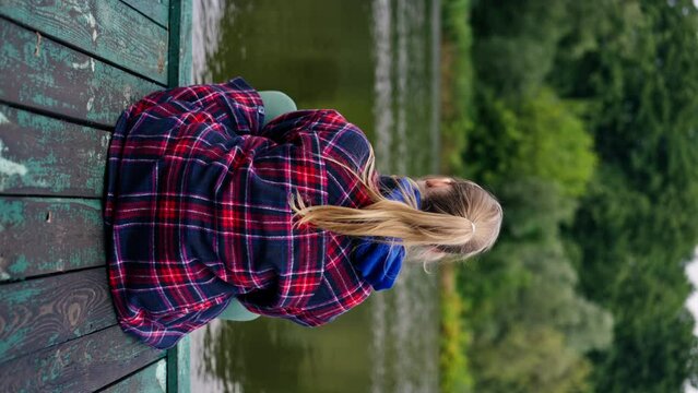 vertical video back view of a girl sitting on the bank of a river or lake in Piers nature depressive state of thoughts
