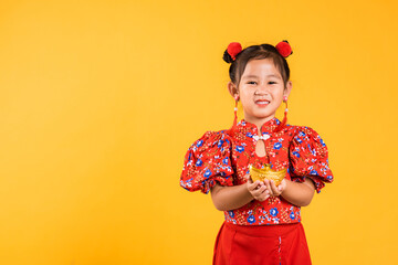 Chinese New Year. Happy Asian Chinese little girl smile wearing red cheongsam holding gold ingot,...
