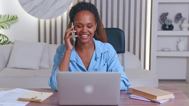 Young happy attractive African American woman student makes phone call to classmate and uses laptop to get online education or write thesis sits at table with books in living room with large sofa.