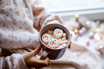 Unrecognizable little child girl holding cup with hot chocolate with marshmallows as snowman. Kid sitting near Christmas decorated window with lights.