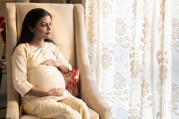 Happy pregnant indian woman touching big belly near window at home. people and expectation concept