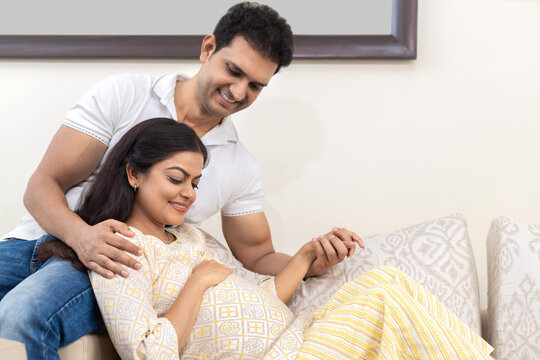 Happy indian couple expecting a baby while husband holding the hand of his pregnant wife sitting on couch. healthcare concept