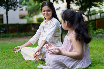 Beautiful young indian mother and her cute daughter doing yoga meditation in the park or garden.