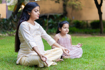 Beautiful young indian mother and her cute daughter doing yoga meditation in the park or garden.