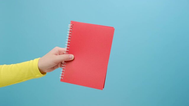 Close up of female hand holding red blank notebook, empty space for commercial text, advertising area on paper, posing isolated over plain blue color background wall in studio. Business idea concept