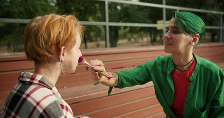 A girl with short green hair in a green shirt makes makeup for her friend with short orange hair in a checkered shirt on brown benches in the park. Lesbian couple of LGBT girls in bright clothes and