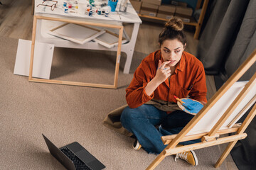 Talented Serious woman professional artist sitting on floor in her studio painting drawing on...