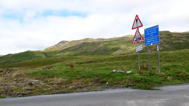 Steep ascent triangle warning road sign in Duddon Valley, white van or minibus driving up the narrow curvy road near Hardknott mountain pass in Lake District National Park, United Kingdom.