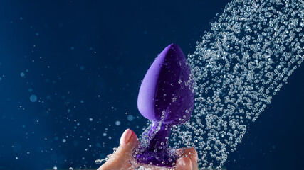 Woman washing purple anal plug under shower on blue background. 
