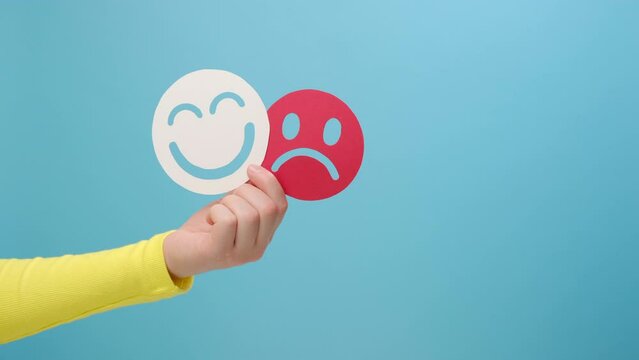 Close up of woman hand holding happy and angry face paper, isolated on blue background. feedback rating, Emotional intelligence, emotion control, mental health assessment, bipolar disorder concept