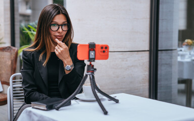 Young elegant hispanic woman in business suit sitting at table at cafe outside streaming via internet using phone on tripod. Beautiful American female chatting with business partner makes video call.