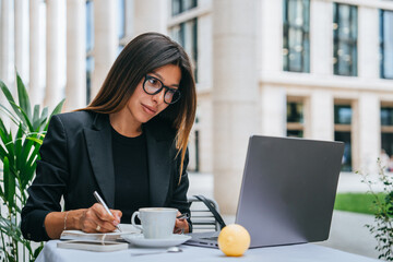 Purposeful brunette young woman in glasses and business suit sitting at cafe outside using laptop...