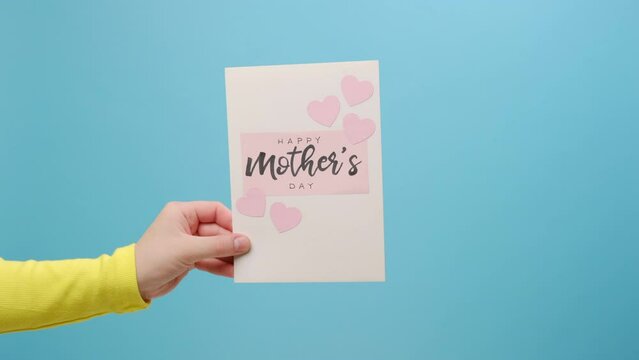 Close up of woman holding card for Mother's Day with little pink hearts, posing isolated on plain blue color background wall in studio with copy space for promotion content. Spring holiday concept