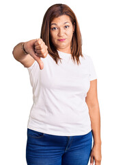 Middle age latin woman wearing casual white tshirt looking unhappy and angry showing rejection and negative with thumbs down gesture. bad expression.