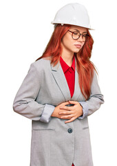Young redhead woman wearing architect hardhat with hand on stomach because indigestion, painful illness feeling unwell. ache concept.