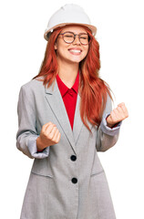 Young redhead woman wearing architect hardhat very happy and excited doing winner gesture with arms raised, smiling and screaming for success. celebration concept.