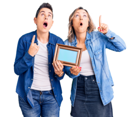 Couple of women holding empty frame amazed and surprised looking up and pointing with fingers and raised arms.