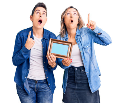 Couple of women holding empty frame amazed and surprised looking up and pointing with fingers and raised arms.