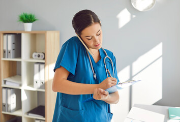 Portrait of a young female doctor or woman nurse standing in office talking by mobile phone and writing notes to clipboard notepad in medical clinic. Medicine and health care concept. - obrazy, fototapety, plakaty