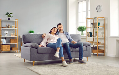Young family couple spending time at home. Happy, relaxed man and woman sitting together on a comfortable sofa in a modern living room interior in their house or apartment - obrazy, fototapety, plakaty