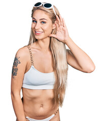Beautiful young blonde woman wearing bikini smiling with hand over ear listening an hearing to rumor or gossip. deafness concept.