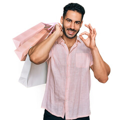 Hispanic man with beard holding shopping bags doing ok sign with fingers, smiling friendly...