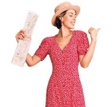 Young beautiful woman wearing summer hat holding map pointing thumb up to the side smiling happy with open mouth