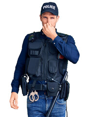 Young handsome man wearing police uniform smelling something stinky and disgusting, intolerable smell, holding breath with fingers on nose. bad smell