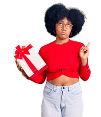 Young african american girl holding gift pointing up looking sad and upset, indicating direction with fingers, unhappy and depressed.