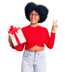 Young african american girl holding gift smiling looking to the camera showing fingers doing victory sign. number two.