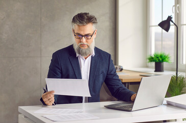 Naklejka premium Mature businessman working with paper document, using laptop in his office. Focused middle aged executive ceo manager in formal wear and glasses analyzing financial data at workplace